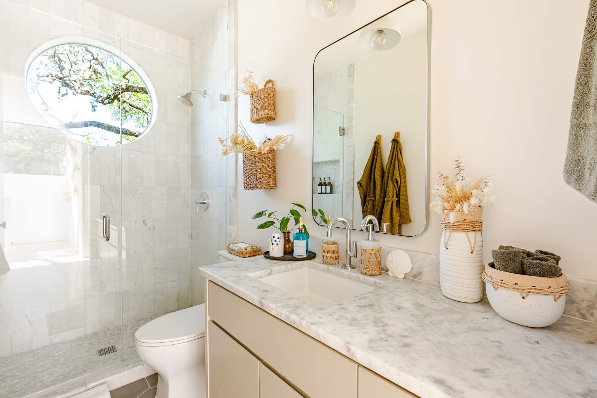 A bathroom with a marble-topped vanity, walk-in glass shower, and a round window looking into the oaks