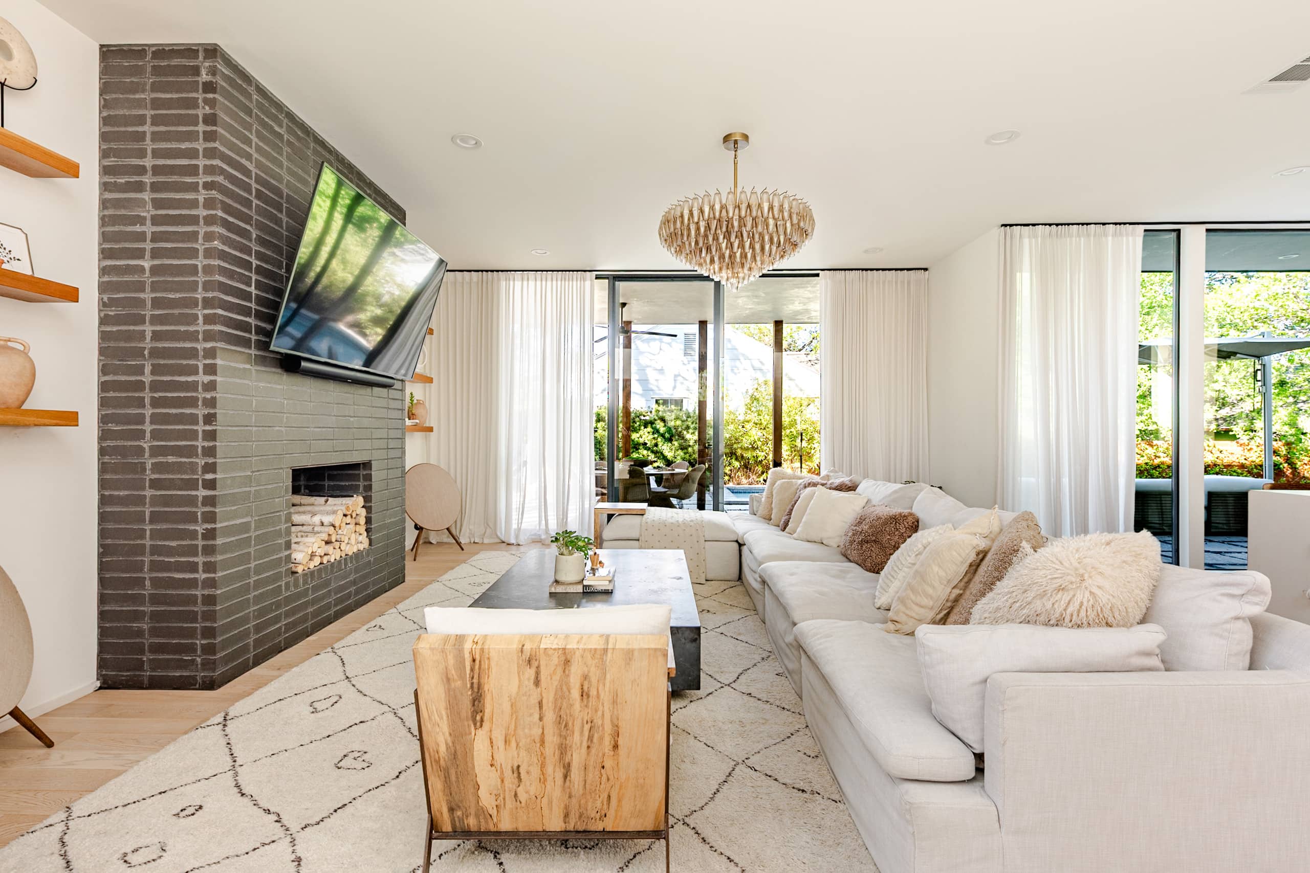 The living room — a plush cream sectional and grey-brick fireplace beneath a cascading brass chandelier, sheer drapes filtering the light