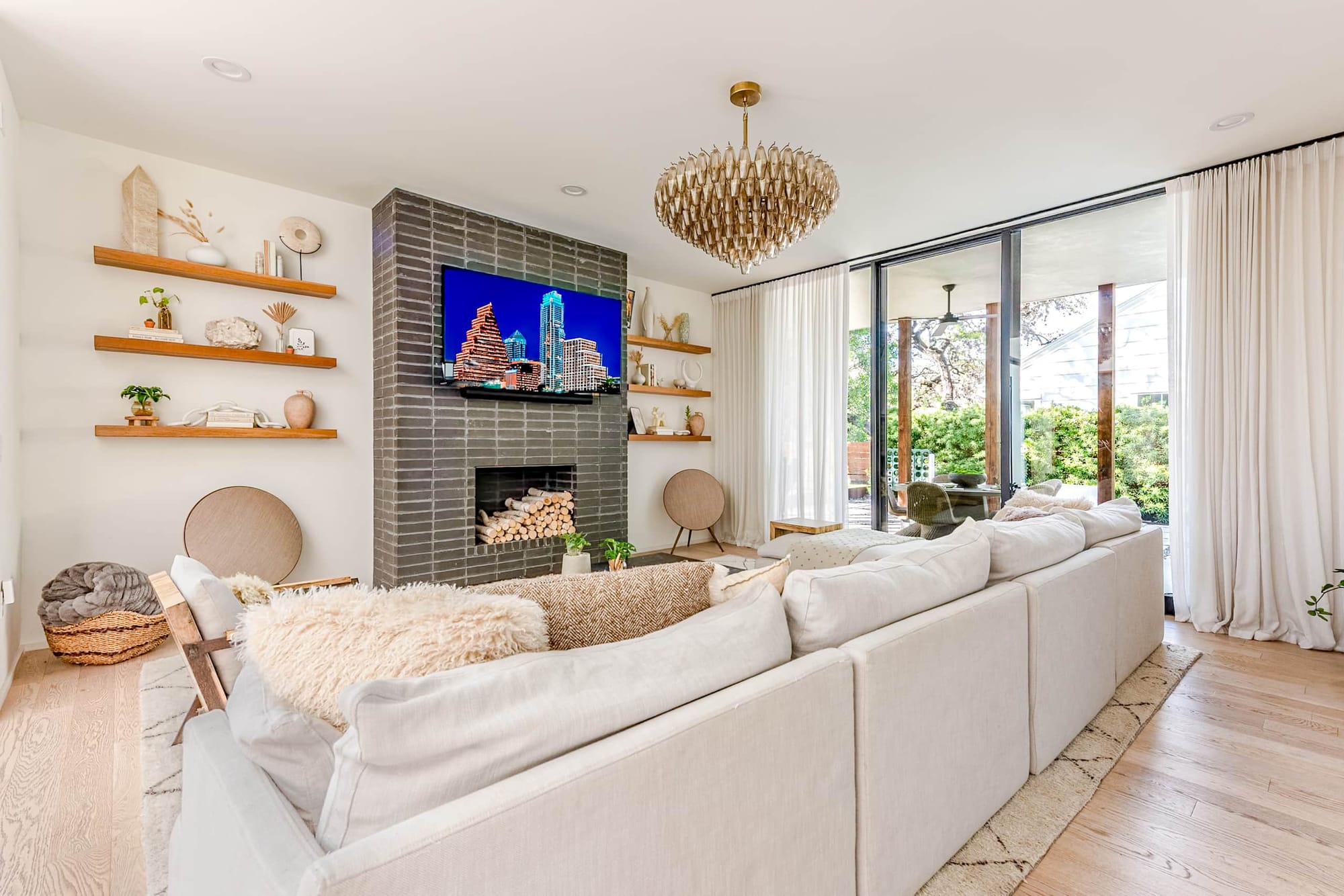 The living room at Bali in Bouldin — a plush cream sectional facing a tile-clad fireplace, floating oak shelves, and a woven brass chandelier in soft natural light