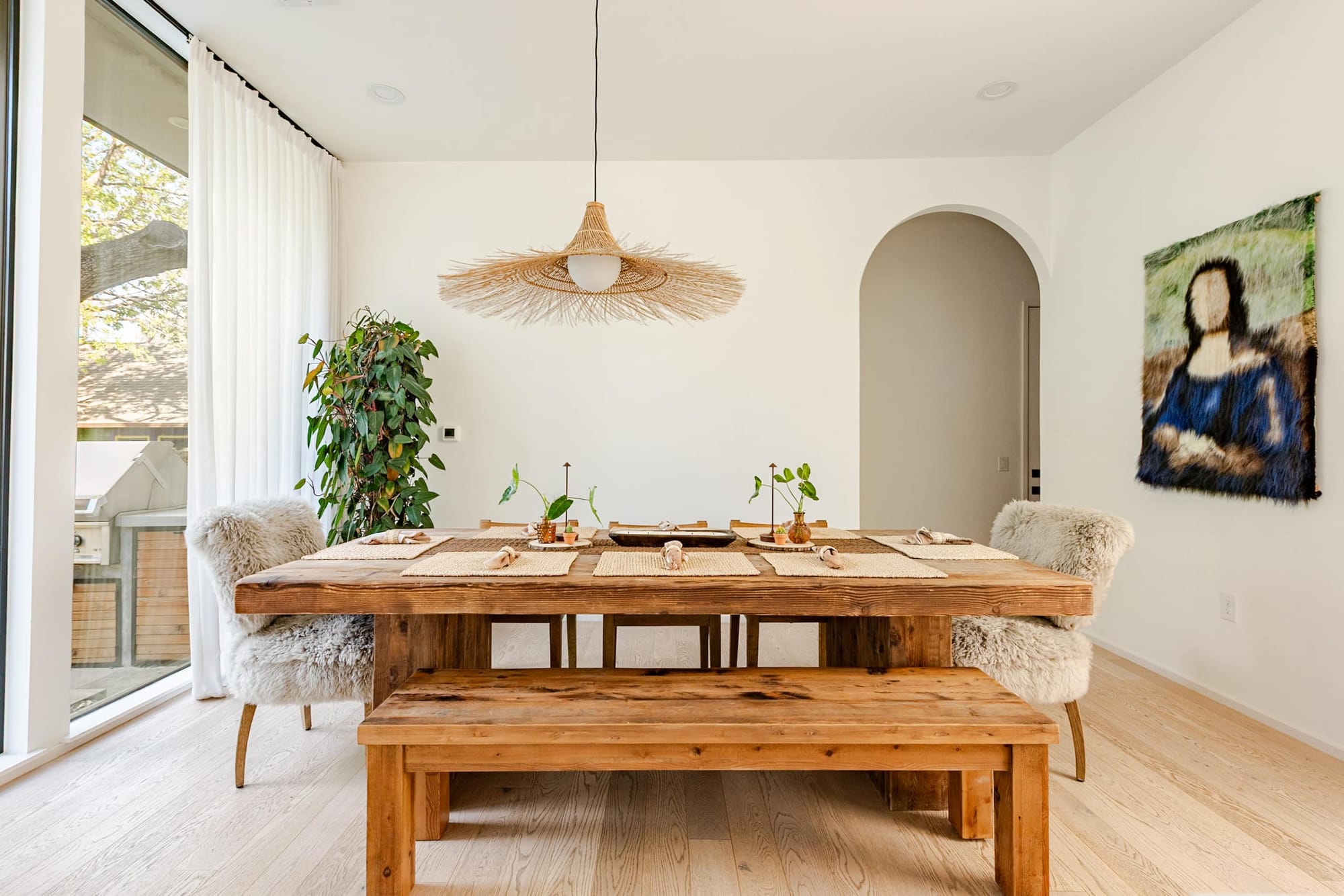 The dining room — a chunky reclaimed-wood table and bench under a sculptural rattan pendant, beside an arched doorway