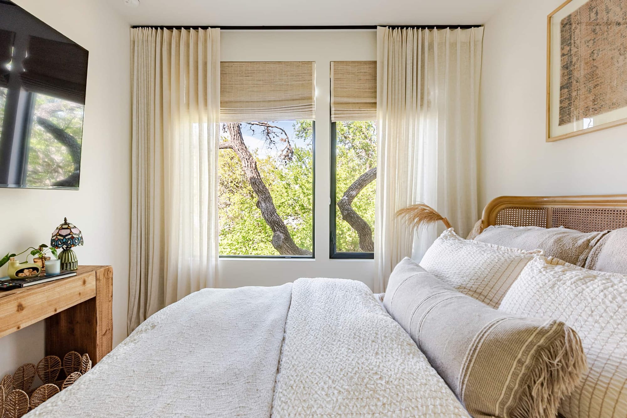 A bedroom with a cane headboard and layered neutral linens, its picture window framing the live-oak canopy